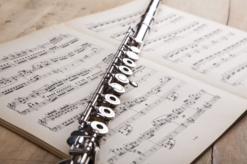 A silver flute lies across an open sheet music book on a light wood surface.