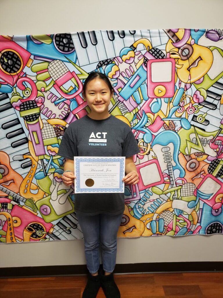 A person stands holding a certificate in front of a colorful, music-themed background. They are wearing a dark grey shirt and blue jeans.