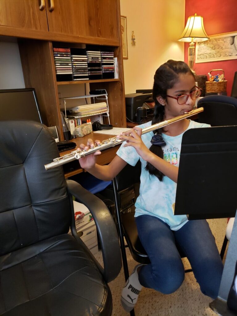 A girl wearing glasses and a blue tie-dye shirt plays the flute while seated in a home office with a music stand in front of her.