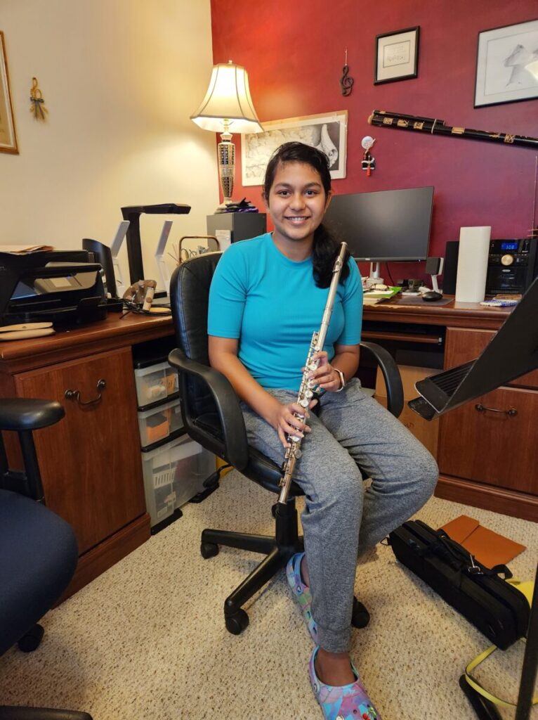A person in a blue shirt and gray pants sits in a home office, holding a musical instrument. Office furniture and equipment surround them.