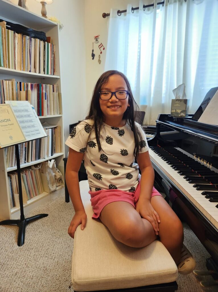 A girl with glasses sits on a bench in front of a piano in a room with shelves full of books and music sheets.