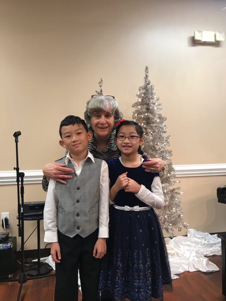An older woman with gray hair hugs a young boy and girl standing in front of a silver Christmas tree in a decorated room.