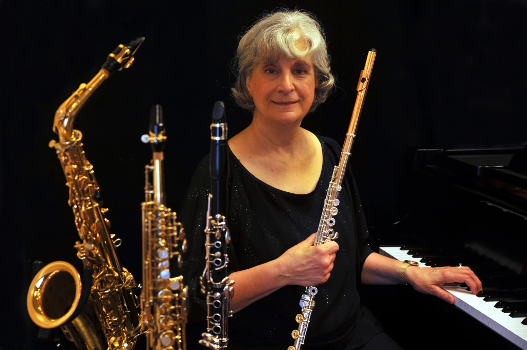 A woman sits at a piano holding a flute, with a saxophone and two clarinets standing beside her against a dark background.